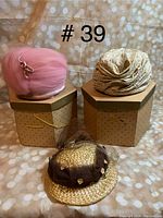 Three vintage hats displayed with two stored on their hexagonal polka dot hat boxes and one straw hat set on the surface in front. Pink hat and gold-toned pleated hat are on boxes. Hats show minor dust from storage but remain in very good condition.