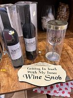 Lot items arranged on wooden surface including two decorative boxed wine bottle-shaped decanters, two bottle-shaped black wine accessory cases containing wine tools, a clear wine glass in plastic holder, and decorative wine sign.