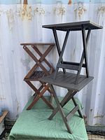 Two wooden folding patio tables stacked and two dark resin folding patio tables stacked on top of them; detail shows slatted surface and cross-brace folding mechanism
