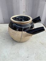 Side view of the ceramic teapot showing the black glaze on the upper half and handle with unglazed beige lower half, no lid.