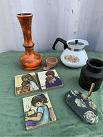 Photo showing entire lot on green cloth backdrop including West German vase, Corning teapot, terracotta piece, black vase, brass and stone holder, and three framed prints.