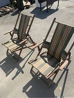 Pair of antique wooden deck chairs viewed from front, showing the striped canvas slings and armrests, placed outdoors on concrete surface.