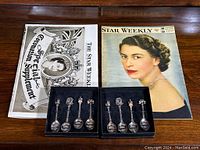 Two vintage newspapers featuring Queen Elizabeth's coronation alongside two black boxes containing a total of eight commemorative silver spoons.