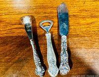 Top view of all three antique sterling silver handled utensils (shoe horn, bottle opener, butter knife) on wooden surface