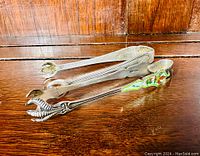 Two antique sterling silver sugar tongs laid on wooden surface, one with enamel decoration, other plain silver.