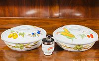 Two Royal Worcester Evesham M round casserole dishes with colorful fruit and vegetable patterns, alongside a small antique egg coddler with metal lid on a wood surface.