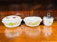 Three porcelain pieces on a wooden shelf: two covered serving dishes with fruit and wheat motif and an egg coddler with metal lid.
