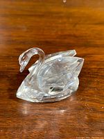 Side angled view of clear Swarovski crystal swan figurine on wooden surface, showing the detailed faceted wings, curved neck, and general shape.