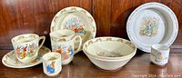 Collection of vintage Bunnykins and Peter Rabbit children's dishes arranged on wooden shelf. Items include cups, bowls, and plates with detailed rabbit illustrations and running rabbit border design.