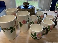 Set of five floral Franciscan mugs with dark branch-like handles, three matching tumblers, and one brown casserole with lid, displayed on a table.