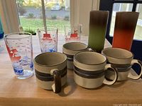 Photo shows 4 ceramic mugs with beige, brown and blue horizontal stripes along with 5 clear glasses decorated with red sailing ships and blue water waves.