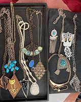 Full view of all 12 necklace and pendant items arranged in a tray, showing variety of metal chains, beads, pendants including turquoise, ivory carved, wooden, and metal designs