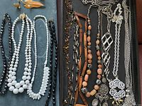 Photo showing a collection of 12 necklaces with white, black, peach, and multicolor beads, along with silver-tone metal pendants and chain elements. The necklaces vary in bead shape and size, including round, faceted, and elongated beads.