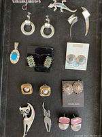 Top view of twelve sterling silver jewelry items showing pins, clip-on earrings, pierced earrings, and a turquoise pendant.