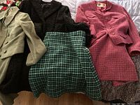 Five wool clothing items spread on a surface: pink Fiandaca suit, black Persian lamb coat, green and navy houndstooth skirt, and additional coats.