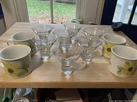 Seven clear footed wine glasses and four ceramic Royal Doulton mugs with floral design arranged on a wooden surface.