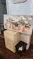 Photo showing the off white metal cabinet, floral canvas art leaning on the wall, and dark wood handcrafted stool in foreground
