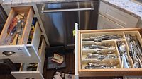 Open kitchen drawers showing various cooking utensils including tongs, whisks, wooden spoons in one drawer; flatware including forks, knives, spoons in an organizer in another drawer.