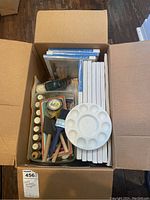 Photo of open cardboard box containing canvases, paintbrushes, paints, palettes, and other supplies.