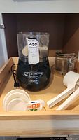 Photo showing black and clear plastic Cuisinart food processor with buttons on front, clear lid and attachment, white plastic potato ricer, and a set of white plastic measuring spoons.