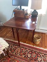 Wooden drop leaf table with polished top surface and drawer beneath, in a room setting with lamp and other furniture, showing overall shape and condition