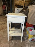 Front view of white wooden end table with single drawer missing pulls, showing scratches and dirt on surface, placed on concrete floor in basement.
