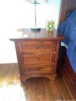 Front view showing one wood end table with three drawers and knob handles, placed on wood floor next to a bed covered in a blue blanket. The table top holds a table lamp and a small decorative item.