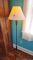 Full view of metal floor lamp with off white shade, standing against teal wall and wood floor.