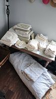 Photo showing folded various white and off white linens and curtains arranged on a wooden table and floor, including sheets, pillowcases, and quilts.