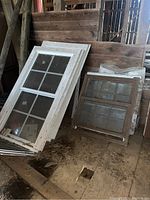 Large and small framed windows leaning against a wall in a wooden structure with visible dust and dirt around.
