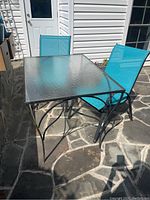 Photo of the rectangular metal frame patio table with textured glass top and one blue mesh chair at an outdoor stone patio