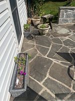 Outdoor patio scene showing two rectangular planter boxes with plants, a cement urn planter, a small wooden bench and a plastic planter on a stone patio.