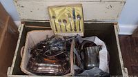 Open trunk containing silver plated serving platters, a pitcher, and boxed flatware set inside.