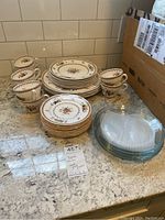 Photo showing Spode china pieces with fruit design and gold trim including teacups, plates in different sizes (dinner, salad, bread dishes), and clear glass plates stacked next to them on countertop.