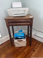 Wooden table with turned legs, single drawer partially open, placed on hardwood floor with printer on top and fabric bins below.
