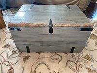 Front view of a rectangular wooden trunk painted grey-blue with heavy wear, showing chipped paint and metal hardware on the corners and front latch.