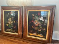 Two floral paintings in wood frames placed against a wall with natural light reflecting on the glass.