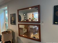 Two matching rectangular wall mirrors with polished wood frames hung vertically on a wall, reflecting room interior including furniture and lighting.