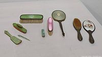 Full set of vintage grooming accessories laid out including hand mirror with floral porcelain back, brass framed hand mirror with beveled glass, two vintage brushes, nail file, lipstick holder, and small green brush.