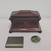 Photo showing antique wooden tea caddy with hinged lid and keyhole, green disc vase with yellow flowers, and Colibri lighter with case.