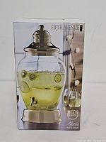 Front view of the Fifth Avenue Crystal Alexis beverage dispenser box showing the product image filled with a yellow beverage and slices of fruit inside. The packaging highlights the 2.5 gallon capacity and gold accents.