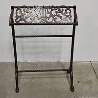 Front view of wooden blanket rack showing carved ornamental top panel with scrollwork and lyre motif, horizontal hanging bars, and claw feet.