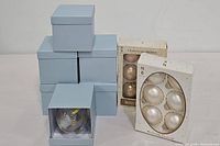 Photo showing 5 blue boxes each containing a candle impression glass ornament with flameless votive inside, and two original boxed sets of Christmas bulbs, one cream colored and one taupe colored, each set having 4 bulbs.