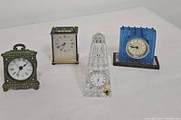 Four vintage clocks on white background, including Waterford crystal obelisk clock, blue glass deco mantel clock, brass carriage clock, and quartz handled clock.