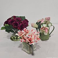 Three floral centerpieces grouped together: red rose bouquet in round glass vase, pink and white amaryllis in square glass vase, pink rolled rose flowers in green watering can-shaped container.