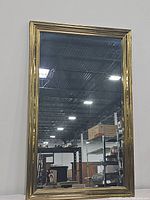 Front view of large vertical rectangular wall mirror with gold ribbed frame showing clear reflection of ceiling and warehouse environment.