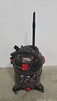 Front angled view of the Hoover wet/dry vacuum showing the black and gray body with red detail, hose wrapped around, and attached upright nozzle.