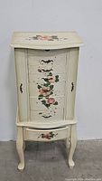 Front view of the cream-colored wooden jewelry armoire with rose floral painted accents and curved legs.