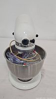 Front-left angled view of the white KitchenAid stand mixer showing the head of the mixer, control dial, and the mixing bowl filled with attachments and power cord.