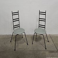 Pair of midcentury metal art accent chairs front and angled view, showing tall black ladder backs with five horizontal bars and gold caps on top. Round light gray padded seats and black metal legs.